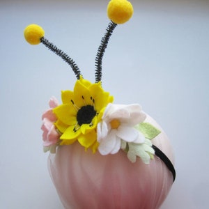 Op de afbeelding: Een zwarte haarband met twee antennes bekroond met gele pompons en een bloemstuk van vilt. De bloemen bestaan uit een centrale felgele zonnebloem, een lichtroze bloesem en een witte margriet met lichtgroene blaadjes.