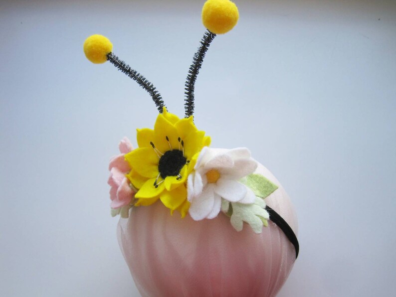 Op de afbeelding: Een zwarte haarband met twee antennes bekroond met gele pompons en een bloemstuk van vilt. De bloemen bestaan uit een centrale felgele zonnebloem, een lichtroze bloesem en een witte margriet met lichtgroene blaadjes.