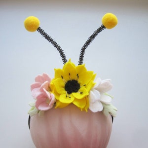 Op de afbeelding: Een haarband met een grote gele viltbloem met een zwart hart, geflankeerd door kleinere roze en witte viltbloemen en lichtgroene blaadjes. Twee zwarte pijpenrager-antennes met gele pompons steken aan de bovenkant uit de haarband.