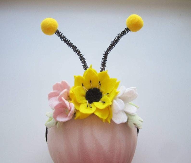 Op de afbeelding: Een haarband met een grote gele viltbloem met een zwart hart, geflankeerd door kleinere roze en witte viltbloemen en lichtgroene blaadjes. Twee zwarte pijpenrager-antennes met gele pompons steken aan de bovenkant uit de haarband.