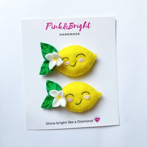 May include: Two yellow felt lemon hair clips attached to a white display card. Each lemon features a smiling face with pink cheeks, a white flower, and two green leaves. The card displays the text Pink & Bright HANDMADE and Shine bright like a Diamond with a small pink diamond icon.