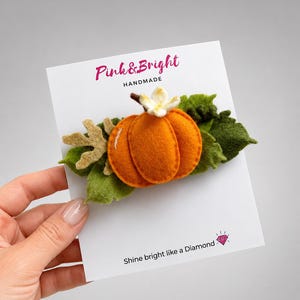 May include: A felt hair clip featuring an orange pumpkin with a small white flower on top, surrounded by green and tan felt leaves. The item is attached to a white display card with the text Pink & Bright HANDMADE and Shine bright like a Diamond.