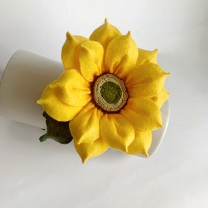 May include: A handcrafted felt sunflower featuring bright yellow petals arranged in multiple layers around a textured, dark green and brown center. A small, dark green felt leaf is attached to the base of the flower, which is resting against a white surface.