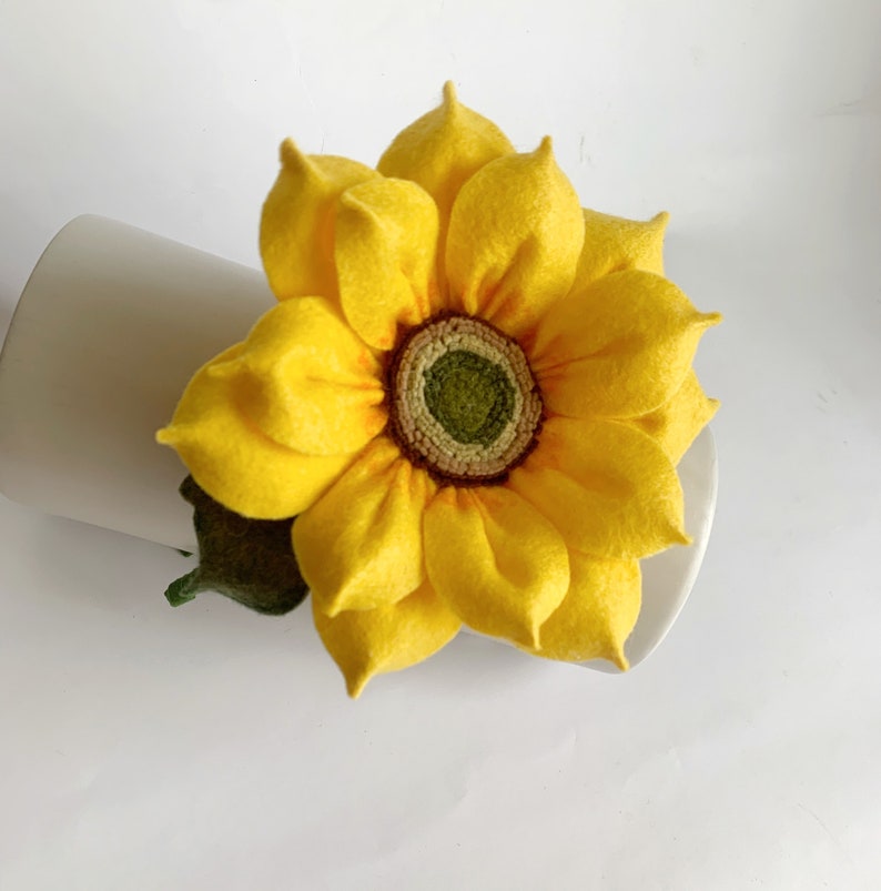 May include: A handcrafted felt sunflower featuring bright yellow petals arranged in multiple layers around a textured, dark green and brown center. A small, dark green felt leaf is attached to the base of the flower, which is resting against a white surface.