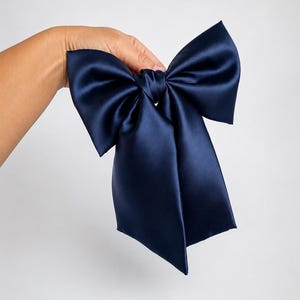 May include: A large, dark navy blue hair bow crafted from a smooth, satin-like fabric. The accessory features two wide, structured loops and two long, flowing tails that extend downward, creating a classic and symmetrical bow shape.