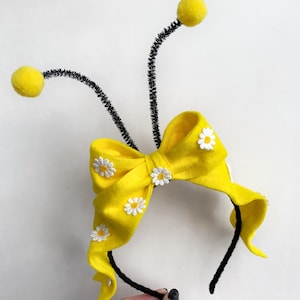 May include: A black headband featuring two tall, flexible black antennae topped with yellow pom-poms. A large, bright yellow fabric bow is attached to the front, decorated with several small, white daisy-shaped floral appliques.