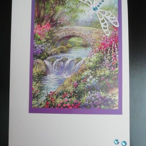 Una hermosa tarjeta hecha a mano de 8x6 que presenta una idílica escena floral junto al río con un puente y una libélula. Decorado con pedrería sintética en color azul.