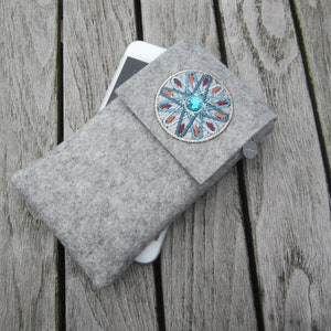 Puede incluir: Una funda para teléfono de fieltro gris con un círculo bordado decorativo que presenta una joya turquesa y detalles dorados.