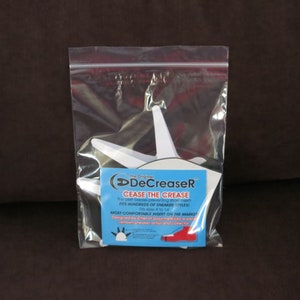 Puede incluir: Un protector de pliegues de zapatos de plástico blanco llamado "DeCreaser" en una bolsa de plástico transparente. El producto está diseñado para evitar pliegues en las zapatillas. El embalaje incluye el texto "The Original DeCreaser", "Cease the Crease", "Fits Hundreds of Sneaker Styles!" y "Most Comfortable Insert on the Market!"