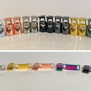 May include: A set of 12 metal shoe lace locks in various colors and finishes, including silver, gold, rose gold, black, and speckled white. The locks are designed to secure shoelaces and add a decorative touch to footwear.