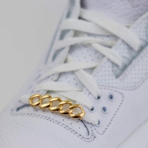 May include: A close-up of a white sneaker with a gold chain lace lock. The chain is made of interlocking links and is a popular accessory for customizing sneakers.