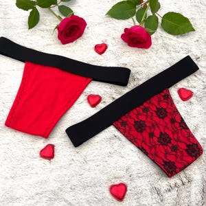 Puede incluir: Dos tangas rojos con cinturillas negras. Uno es rojo liso, el otro es de encaje rojo con un estampado floral. Caramelos rojos en forma de corazón y rosas rojas están dispersos.