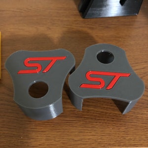 Op de afbeelding: Twee grijze plastic auto-emblemen met de letters "ST" in rood. De emblemen zijn ontworpen om aan een auto te worden bevestigd.