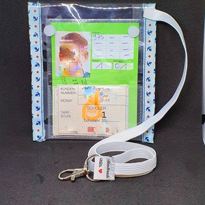 Könnte beinhalten: Ein durchsichtiger Plastik-Ausweishalter mit einem weißen Lanyard und einem silbernen Clip. Der Halter hat eine grüne Karte mit dem Text "975" und ein blau-weiß gestreiftes Band. Das Lanyard hat ein weißes Etikett mit dem Text "Handgefertigt mit Liebe".