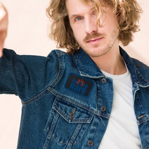 Könnte beinhalten: Ein Mann trägt eine blaue Jeansjacke mit einem schwarzen Patch, auf dem "Raising Rare" in weißer Schrift mit einem roten Symbol von drei Personen steht.
