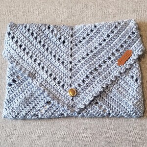 May include: A light blue crocheted clutch with a fold-over flap and a gold-colored button closure. The handmade bag features a textured, open-weave design and a small brown leather tag that reads "Handmade".