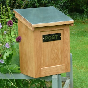 May include: A wooden post box with a metal top and a brass "POST" sign. The post box is mounted on a fence post.