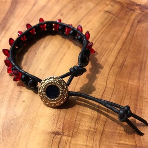 May include: A black leather bracelet with red and black beads and a gold button closure.