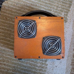 Puede incluir: Una caja de madera con dos cubiertas de ventilador metálicas circulares. La caja tiene un asa negra y está hecha de madera marrón claro. Los ventiladores están cubiertos con una malla de alambre plateada. La caja está sobre una alfombra estampada.