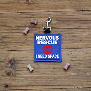 May include: A blue dog tag with the text "NERVOUS RESCUE STOP I NEED SPACE" in white lettering and a red stop sign. The tag is attached to a silver metal clip. Several dog treats are scattered around the tag on a wooden surface.