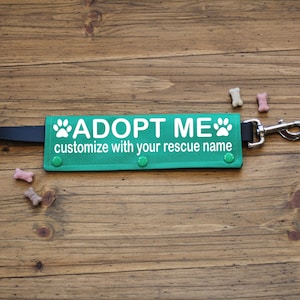 May include: A green dog leash attachment with the text "ADOPT ME" and "customize with your rescue name" in white. The attachment has a silver clasp and is decorated with paw prints. Several dog treats are scattered around the leash.