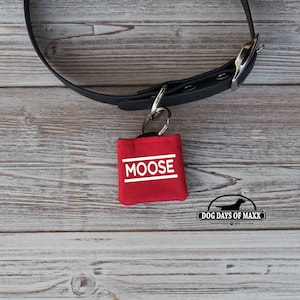 May include: A red dog tag holder with the name "MOOSE" in white block letters. The tag is attached to a black dog collar with a silver metal clasp. The Dog Days of Maxx logo is in the bottom right corner.