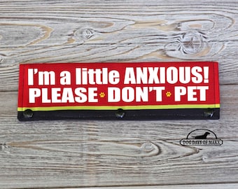 May include: Red and black dog training aid with the text "I'm a little ANXIOUS! PLEASE DON'T PET" and a paw print. The text is in white and the paw print is yellow. The aid is on a wood background. The bottom right corner has a logo that says "DOG DAYS OF MAXX".