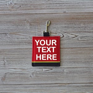 May include: Red fabric keychain with a black clip and a white embroidered message that says "YOUR TEXT HERE".