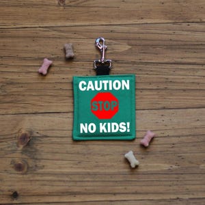 May include: A green dog leash holder with the text "CAUTION STOP NO KIDS!" in white. A red stop sign is in the center. The holder has a silver metal clip. Several dog treats are scattered around the holder on a wooden surface.