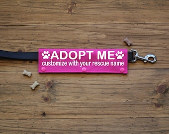 Op de afbeelding: Een felroze hondenriem bevestiging met de witte woorden "ADOPT ME" en pootafdrukken. De tekst "customize with your rescue name" is ook opgenomen. De riem is zwart en de hardware is zilver.