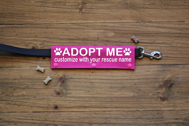 May include: A bright pink dog leash attachment with the words "ADOPT ME" in white, along with paw prints. The text "customize with your rescue name" is also included. The leash is black, and the hardware is silver.