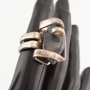 May include: A silver ring with a black stone set in a geometric design. The ring is on a black mannequin hand.