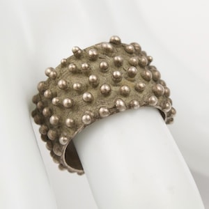 May include: A wide silver-toned ring featuring a textured, matte surface covered in numerous small, raised spherical studs. The metal has a weathered, oxidized appearance with a consistent pattern of bumps across the entire outer band.