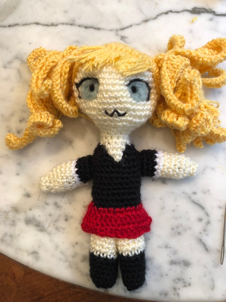 May include: A crocheted doll with yellow hair, a black and white dress, and a red skirt. The doll has blue eyes and a white face.