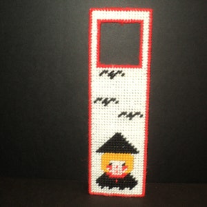 May include: A white and red plastic bookmark with a black and yellow witch design. The witch is wearing a black hat and a black dress. The bookmark has a rectangular cutout in the top for a page.