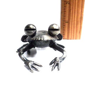 May include: A small, silver metal frog sculpture with large, round eyes. The frog is sitting on a white surface.