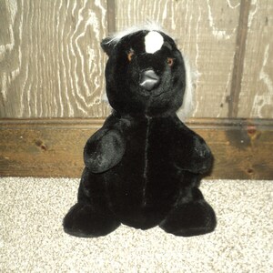 May include: Black plush skunk toy with a white stripe on its forehead. The toy has a black nose and small black eyes.