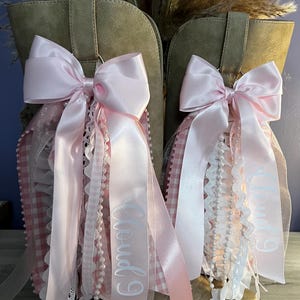 May include: A pair of tan suede boots adorned with decorative pink satin bows. Each bow features cascading ribbons, white lace trim, pearl-like beaded strands, and pink gingham fabric. The text "Cloud 9" is printed in white on the long satin ribbons.