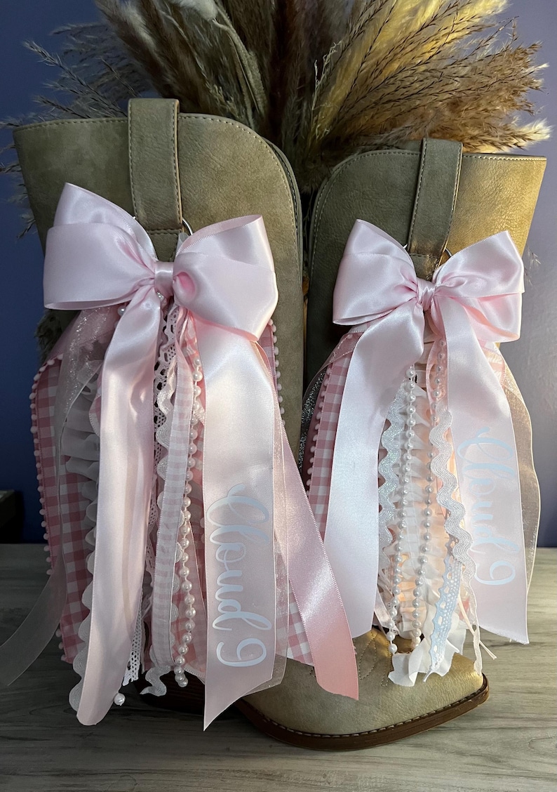 May include: A pair of tan suede boots adorned with decorative pink satin bows. Each bow features cascading ribbons, white lace trim, pearl-like beaded strands, and pink gingham fabric. The text "Cloud 9" is printed in white on the long satin ribbons.