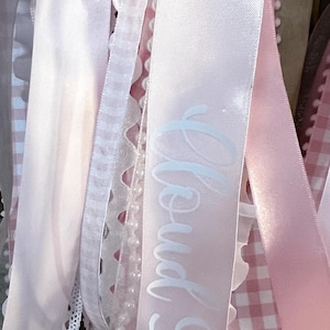 May include: A collection of hanging ribbons in various shades of light pink and white. The assortment includes satin ribbons, lace trim, a string of small white faux pearls, and a pink and white gingham ribbon. One satin ribbon features the text Cloud 9.