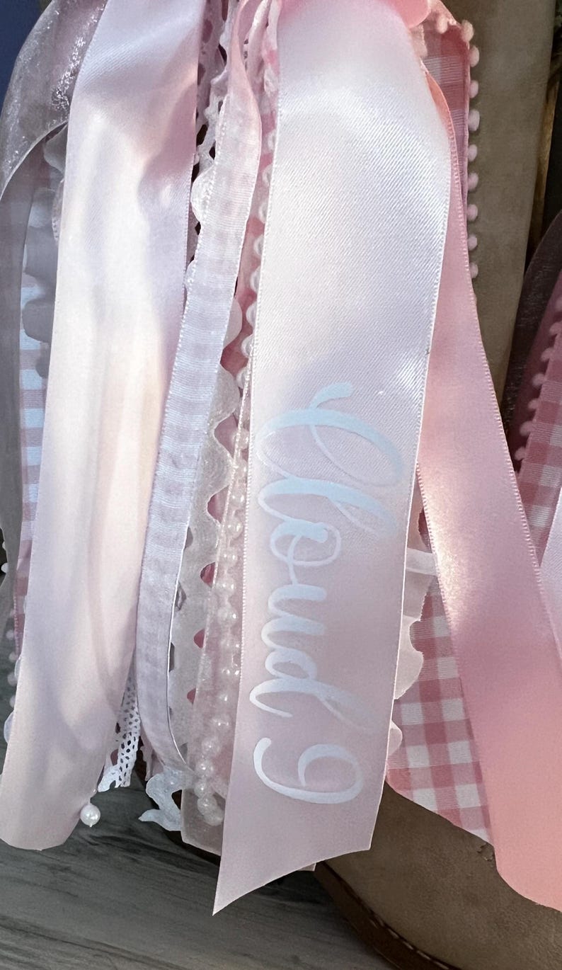 May include: A collection of hanging ribbons in various shades of light pink and white. The assortment includes satin ribbons, lace trim, a string of small white faux pearls, and a pink and white gingham ribbon. One satin ribbon features the text Cloud 9.