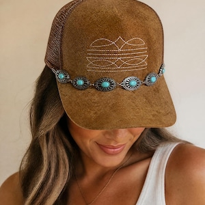May include: A brown suede trucker hat featuring a mesh back and a structured front panel. The front is decorated with white stitched geometric embroidery and a horizontal silver-toned metal chain adorned with oval turquoise-colored cabochon stones.
