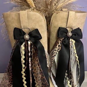 May include: A pair of tan boots adorned with decorative black satin bows. Each bow features a central lion head emblem and is accented with hanging ribbons in leopard print, black lace, gold rickrack trim, and shimmering sequins.