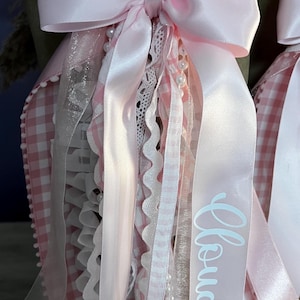 May include: A decorative accessory featuring a large light pink satin bow with cascading ribbons, white lace trim, and a string of small white pearls. One ribbon displays the text Cloud 9 in white script. The base is a light pink and white gingham fabric.