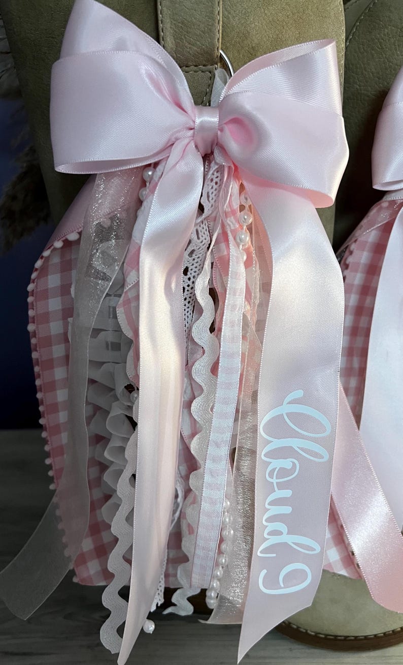 May include: A decorative accessory featuring a large light pink satin bow with cascading ribbons, white lace trim, and a string of small white pearls. One ribbon displays the text Cloud 9 in white script. The base is a light pink and white gingham fabric.