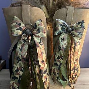 May include: A pair of tan boots adorned with decorative bows featuring a camouflage pattern. Each bow is centered with a dark, textured circular button and includes cascading ribbons in shades of olive green, black, and metallic gold sequins.