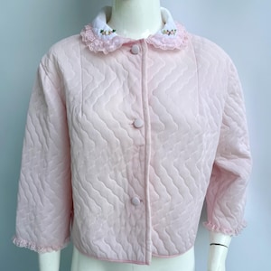 Puede incluir: Chaqueta acolchada rosa claro con cuello blanco y bordado floral. La chaqueta tiene cierre de botones, mangas tres cuartos y ribete de encaje festoneado. La chaqueta se muestra en un maniquí.