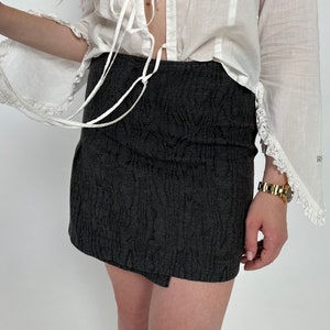 May include: A black mini skirt with a textured pattern. The skirt has a wrap style and a side slit.