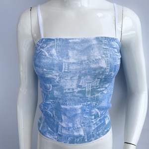 May include: A light blue denim-print tube top with thin white straps. The top is displayed on a white mannequin. The fabric has a faded, textured appearance, mimicking the look of denim jeans.