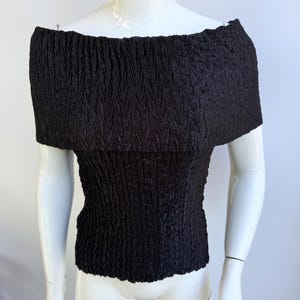 Op de afbeelding: Zwarte off-the-shoulder top van getextureerde, gekreukelde stof. De top heeft een getailleerde taille en een brede, gevouwen halslijn. De stof heeft een subtiel, verticaal patroon. Het kledingstuk wordt getoond op een witte mannequin.